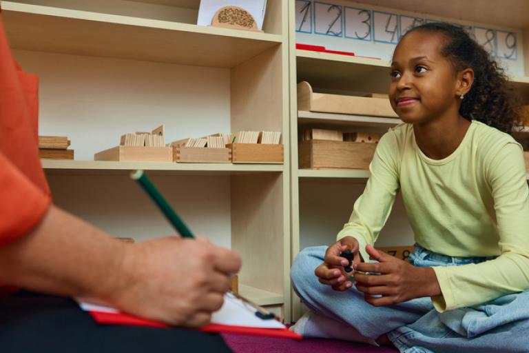 Image of a child sitting cross legged on the floor listening to an adult holding a clipboard and pencil