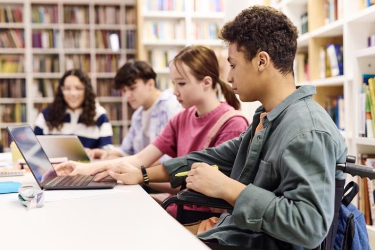 Group of 4 teenagers sitting at a desk in a library working on laptops